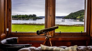 A room with a large window offering a view of a river with boats, a grassy area, and hills. A telescope is placed on a tripod in the foreground. - Shieldaig Lodge Hotel (Gairloch)