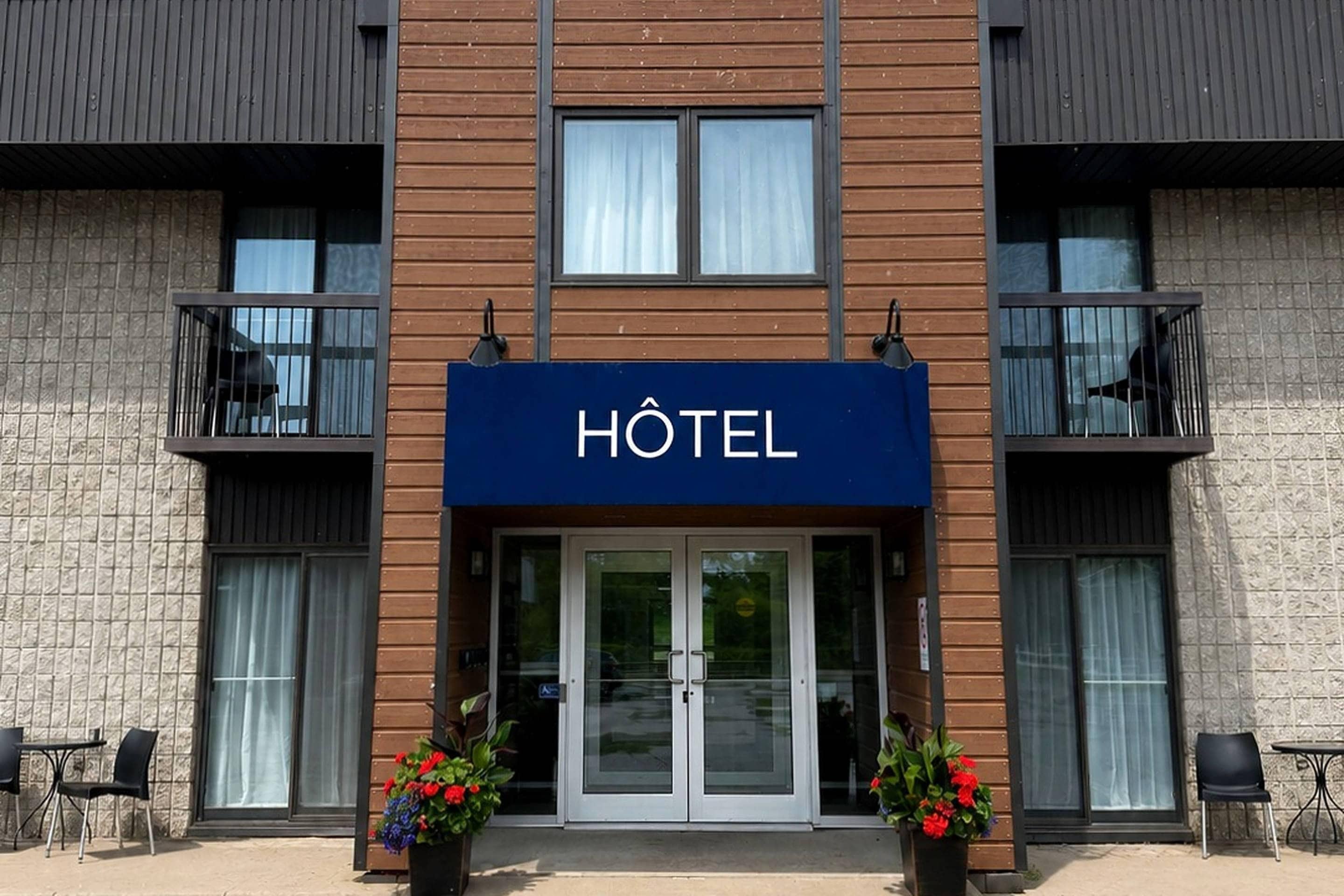 Ein Hotel Eingang mit einem Schild mit der Aufschrift 'HÔTEL' über den Türen, zwei Blumentöpfe mit Blumen und eine kleine Außen-Sitzgelegenheit mit Stühlen.
