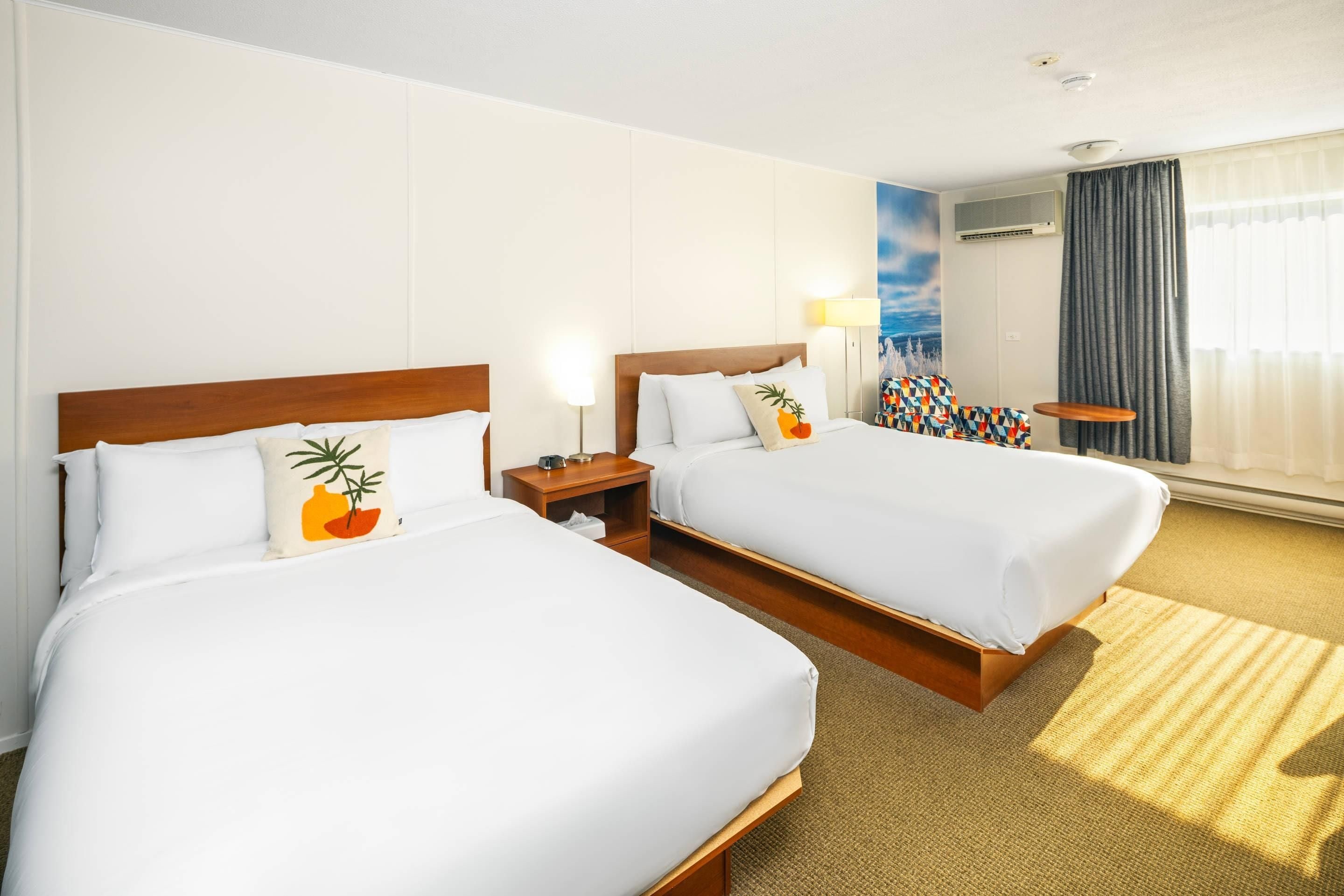 Ein Hotelzimmer mit zwei Betten, einem Nachttisch, einer Lampe, einem Fenster mit Vorhängen und einem Wandbild.