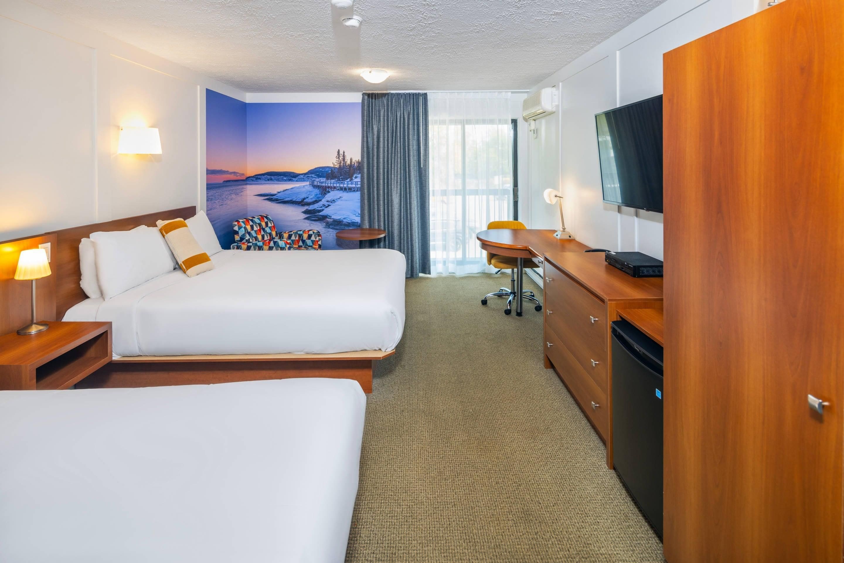Habitación de hotel con dos camas, un escritorio, un televisor y un amplio ventanal con vistas panorámicas.