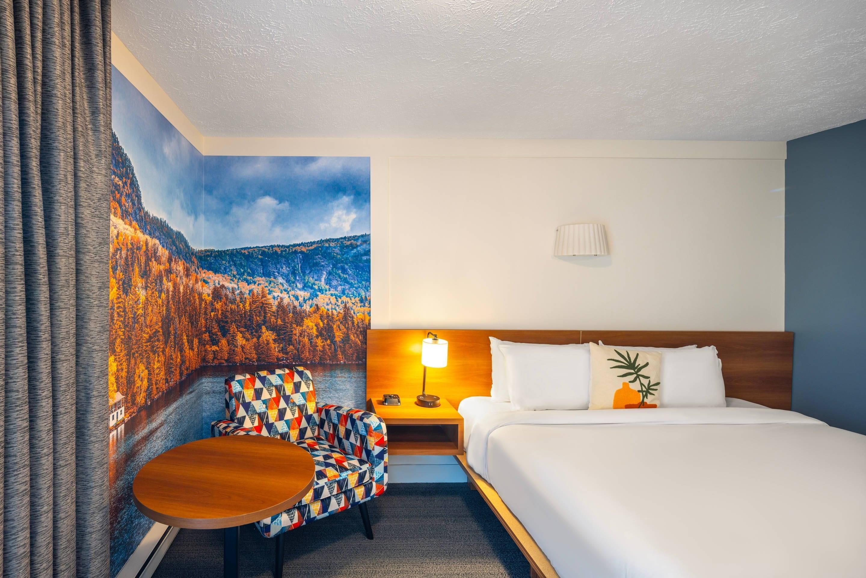 Une chambre d’hôtel avec un lit, une table de chevet, une lampe, une chaise et un tableau représentant un paysage forestier.