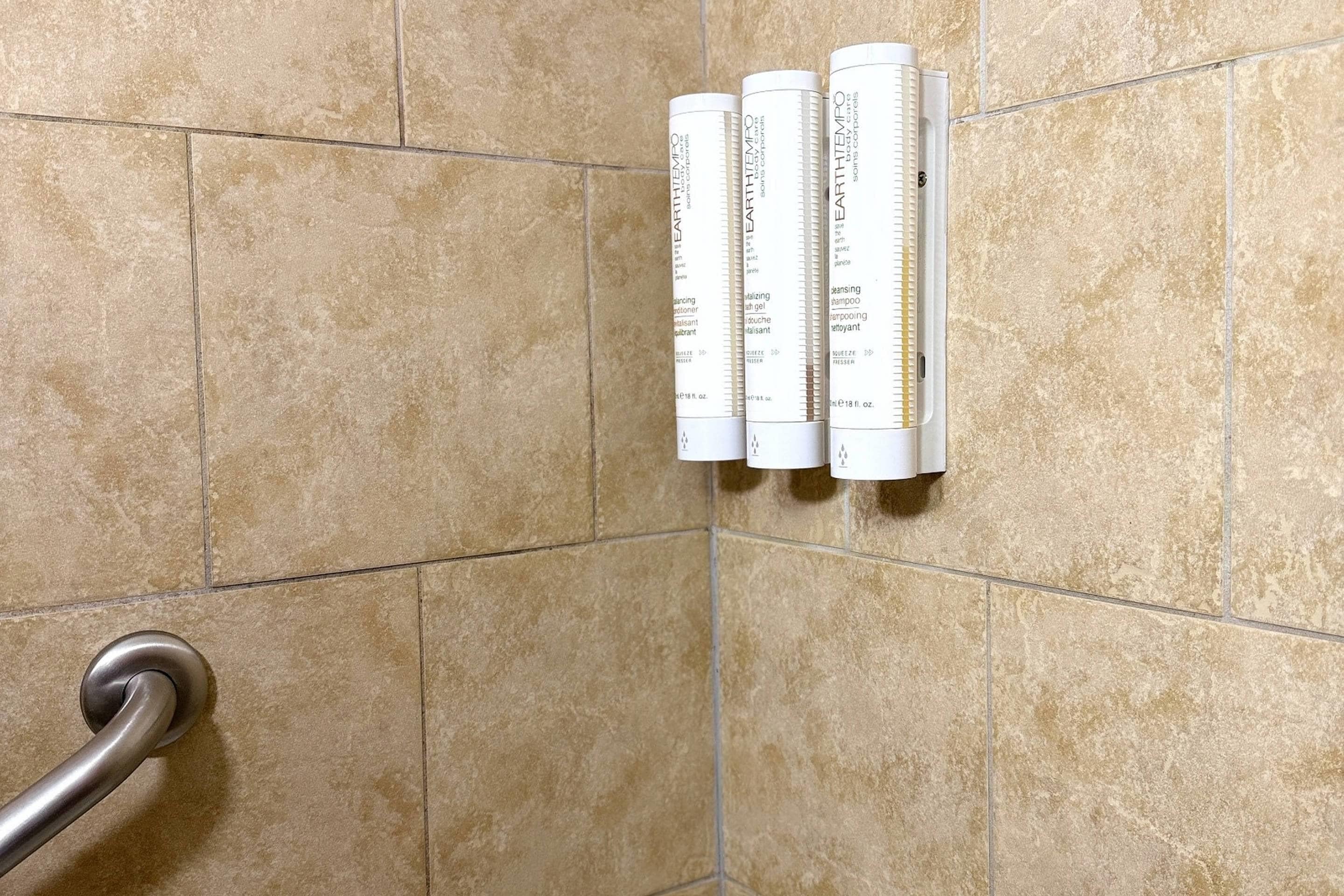 Une salle de bain avec des carreaux de couleur beige, une barre d’appui et trois bouteilles blanches étiquetées « EARTHTEMPO » sur un mur.