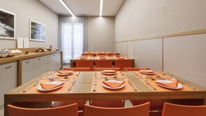 Een eetgedeelte met oranje stoelen en tafels, voorzien van bestek en borden, plus een keuken met kasten en een aanrecht.