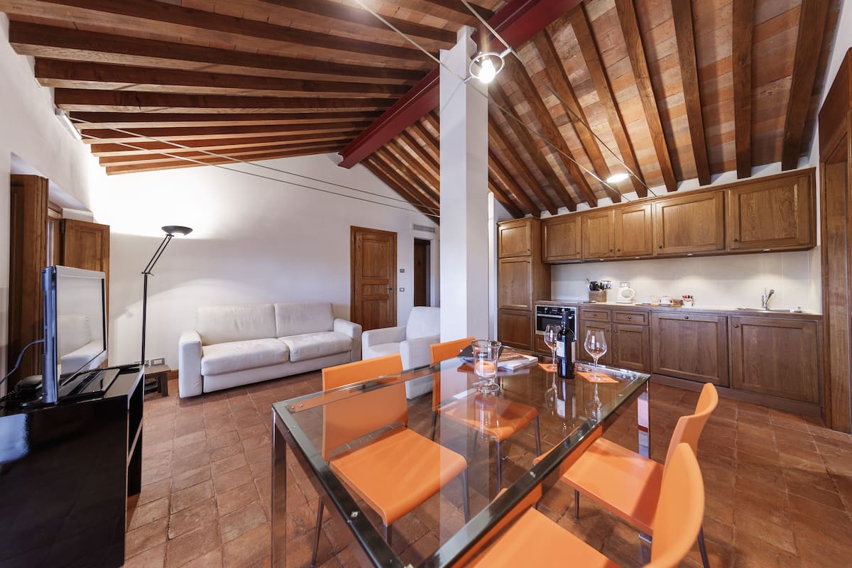 Una cocina moderna con armarios de madera, un comedor con mesa de vidrio y sillas naranjas, y un sofá blanco en la sala de estar.