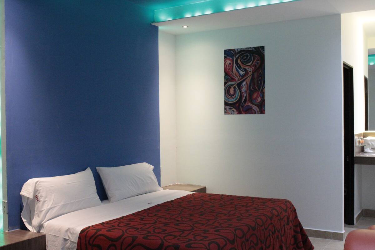 Habitación de hotel con cama, mesita de noche, un cuadro en la pared y baño a la vista a través de una puerta abierta.