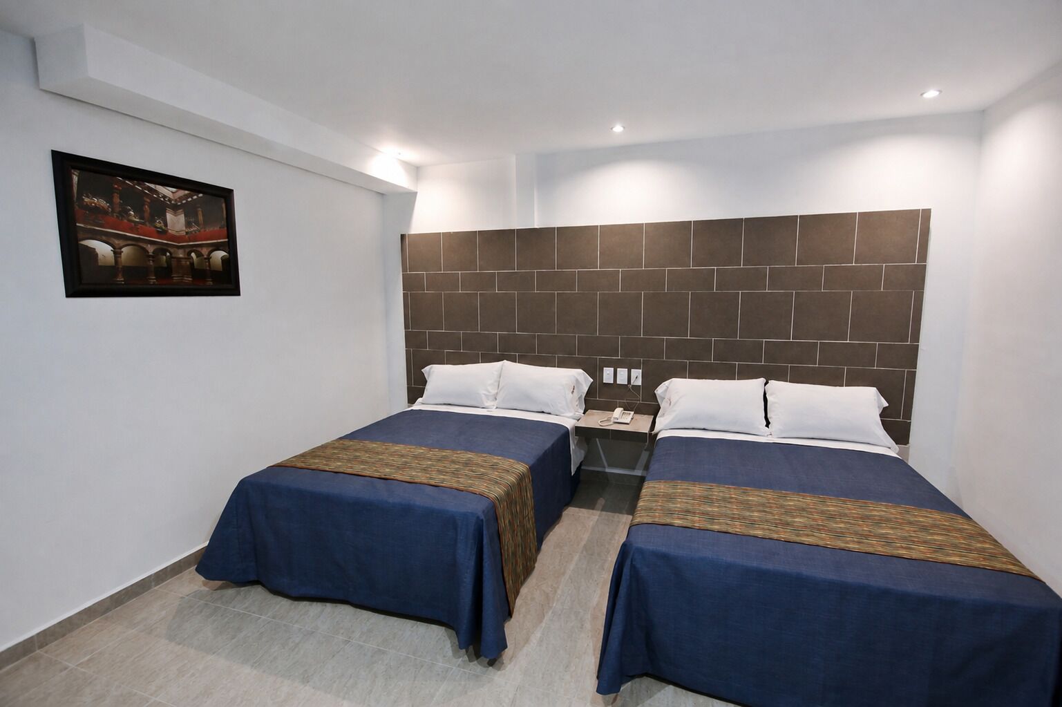 A hotel room with two beds, a headboard with a brick pattern, a small bedside table with a phone, and a framed picture on the wall.