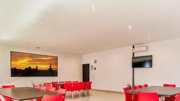 Un salón de conferencias con sillas y mesas rojas, un cuadro grande de un atardecer y un televisor de pantalla plana en la pared.