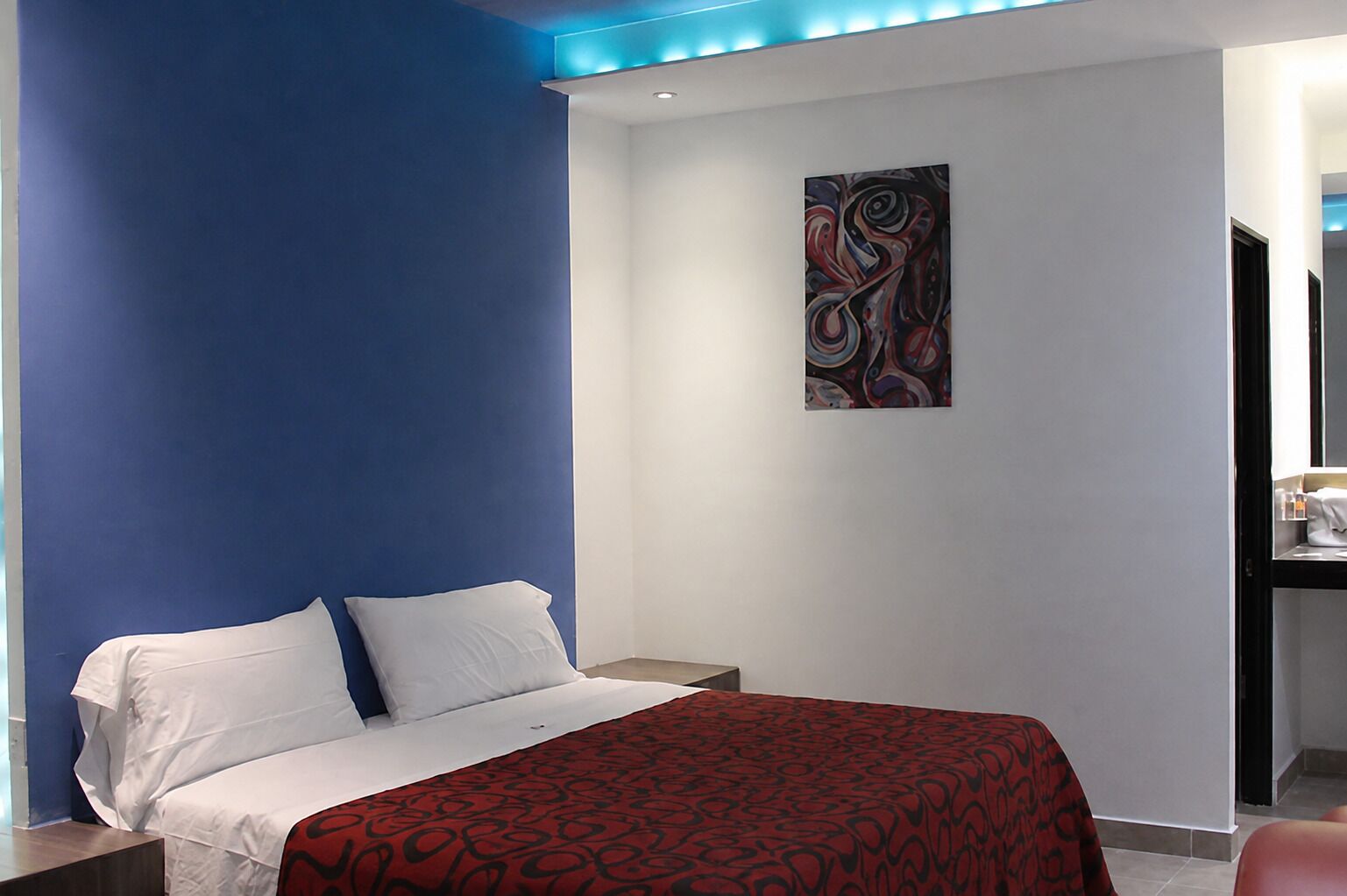 A hotel room with a bed, a nightstand, a desk with a chair, and a painting on the wall.