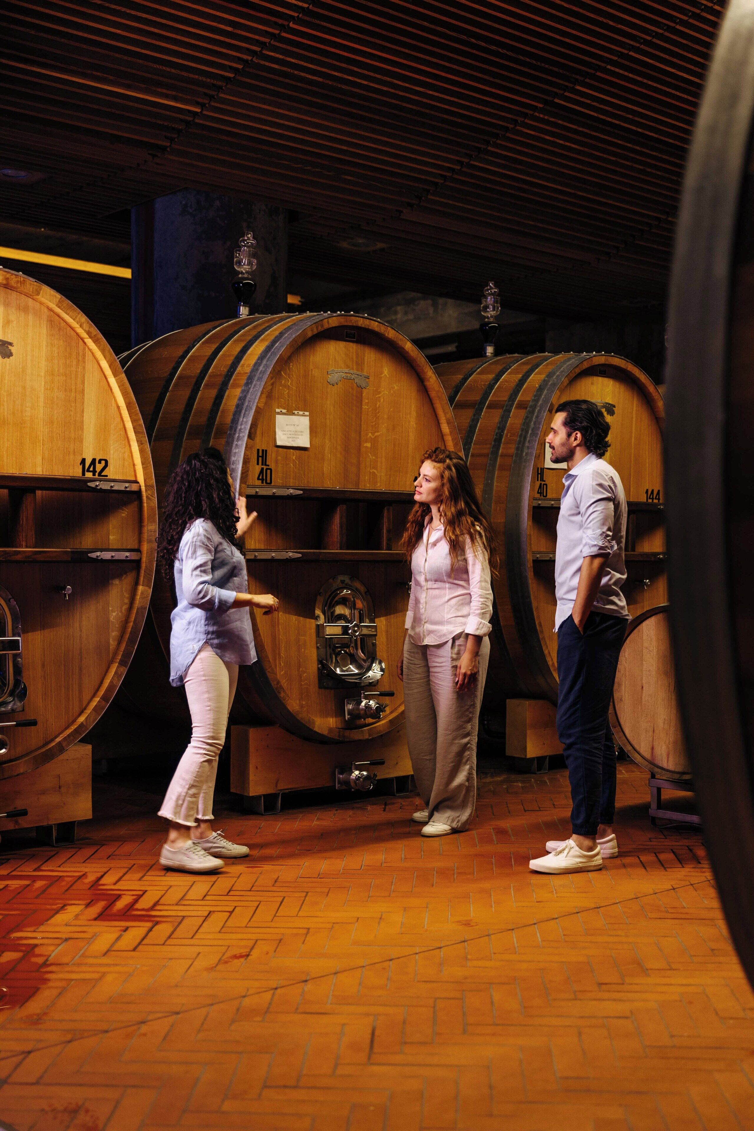 Three people standing in a cellar with wooden barrels, each barrel has a number on it.