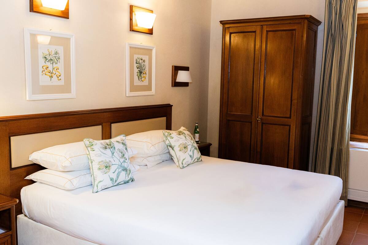 Een opgemaakt bed met bloemenkussens, een houten garderobe en ingelijste botanische prints aan de muur.