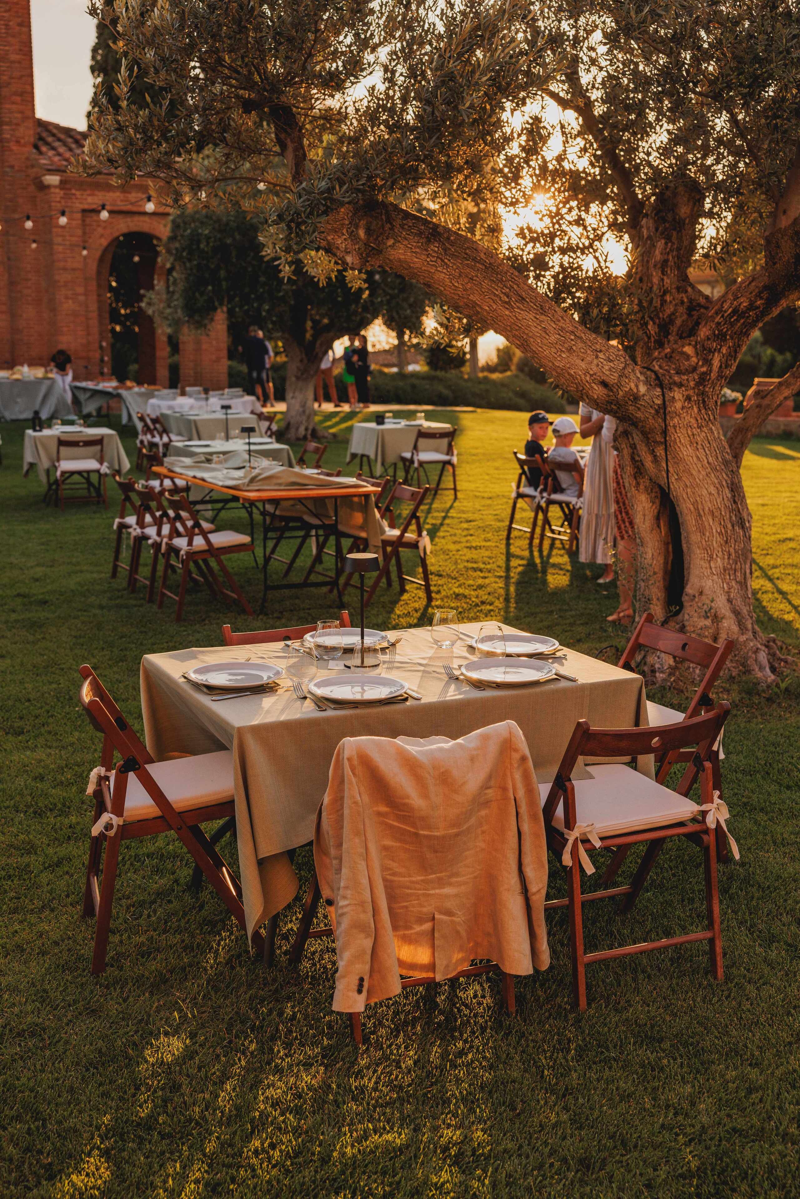 Un área de comedor al aire libre con mesas dispuestas para una comida, rodeada de sillas y un árbol grande.