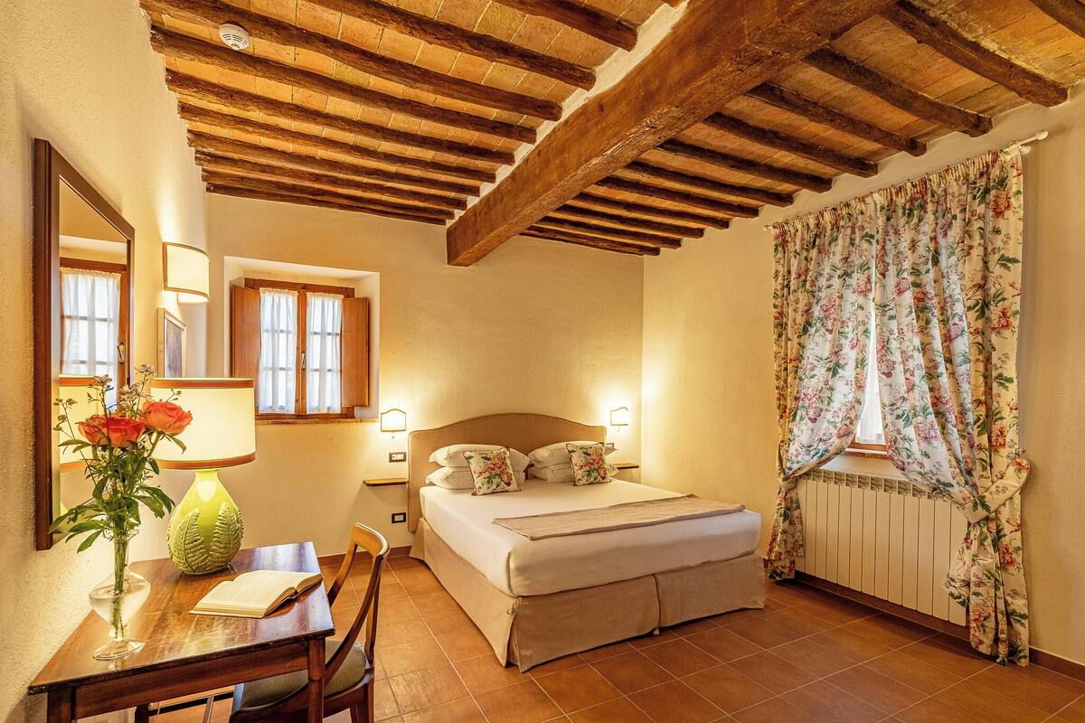 Een slaapkamer met een houten plafond, een bed met bloemen gordijnen, een nachtkastje met een lamp en een bloemenvaas, en een raam met gordijnen.
