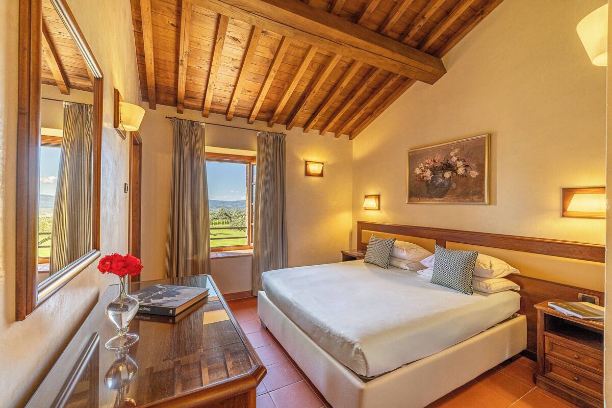 Een slaapkamer met een groot bed, houten balken in het plafond, een nachtkastje, een bijzettafeltje met een boek en een bloevas, en een schilderij aan de muur.