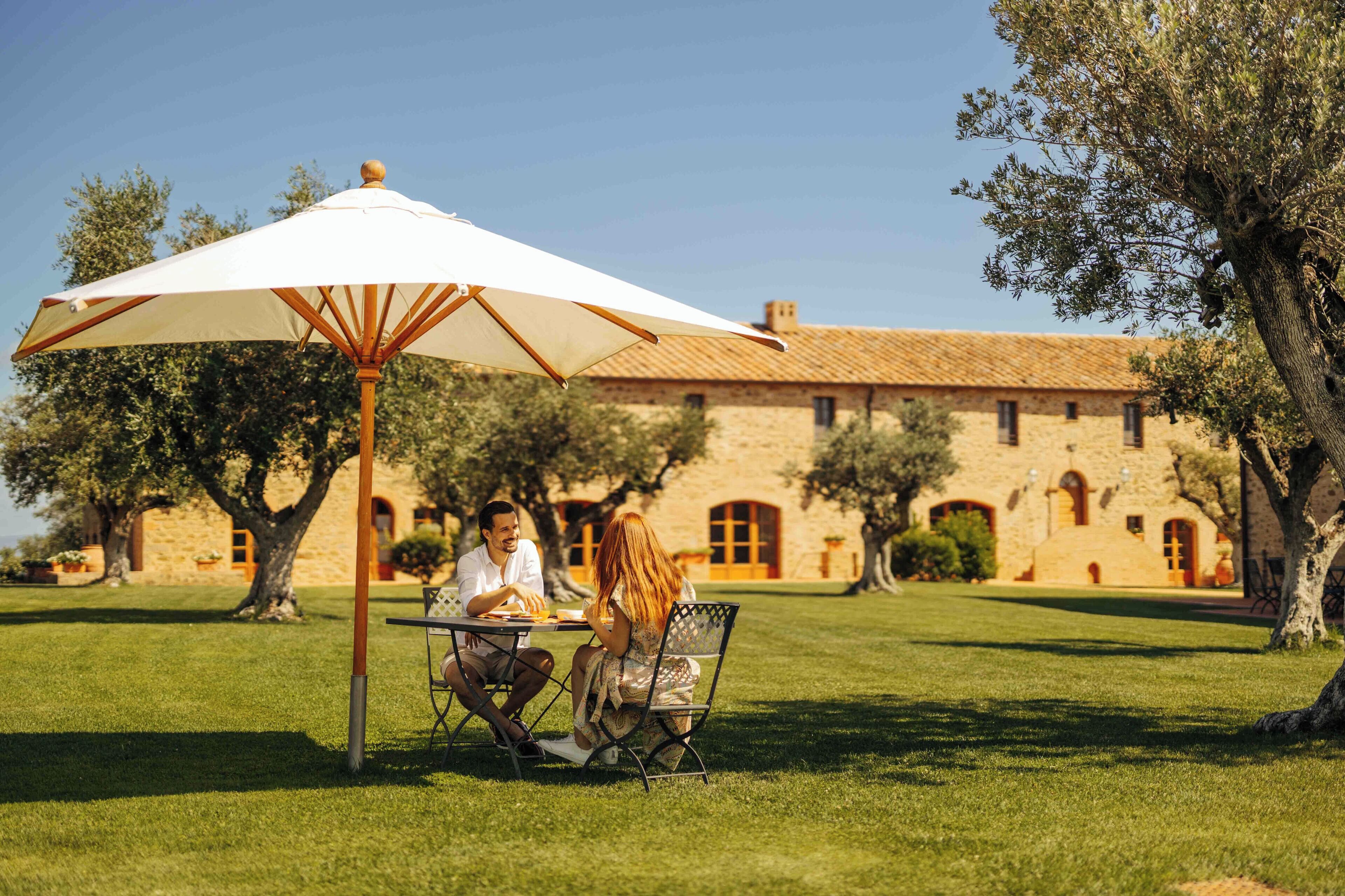 Un couple est assis à une table, dînant sous un parasol blanc, dans un espace extérieur verdoyant, avec un bâtiment en pierre en arrière-plan.