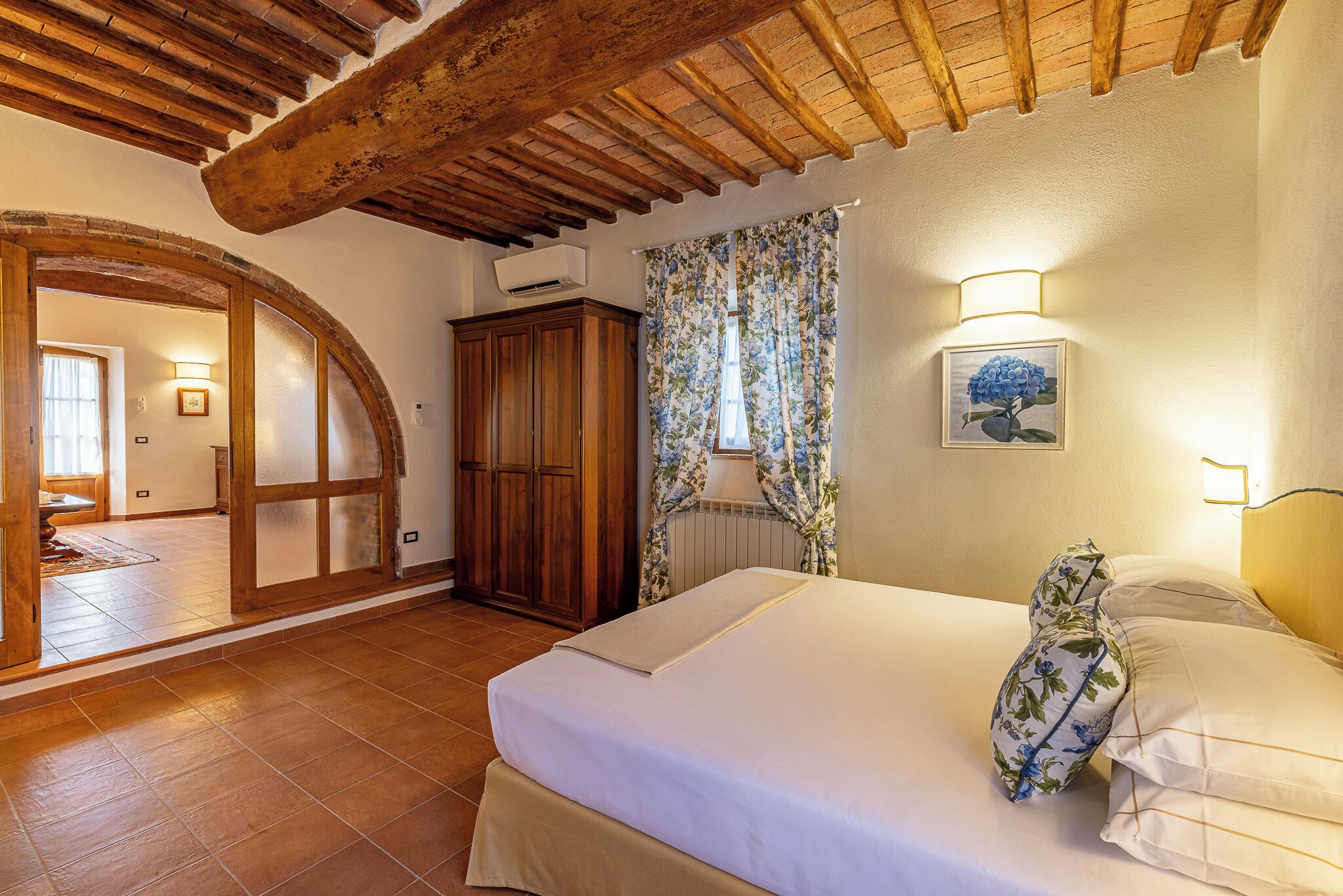 Una camera da letto con soffitto in legno, un letto con biancheria bianca, un cuscino a motivi floreali e una finestra con tende.
