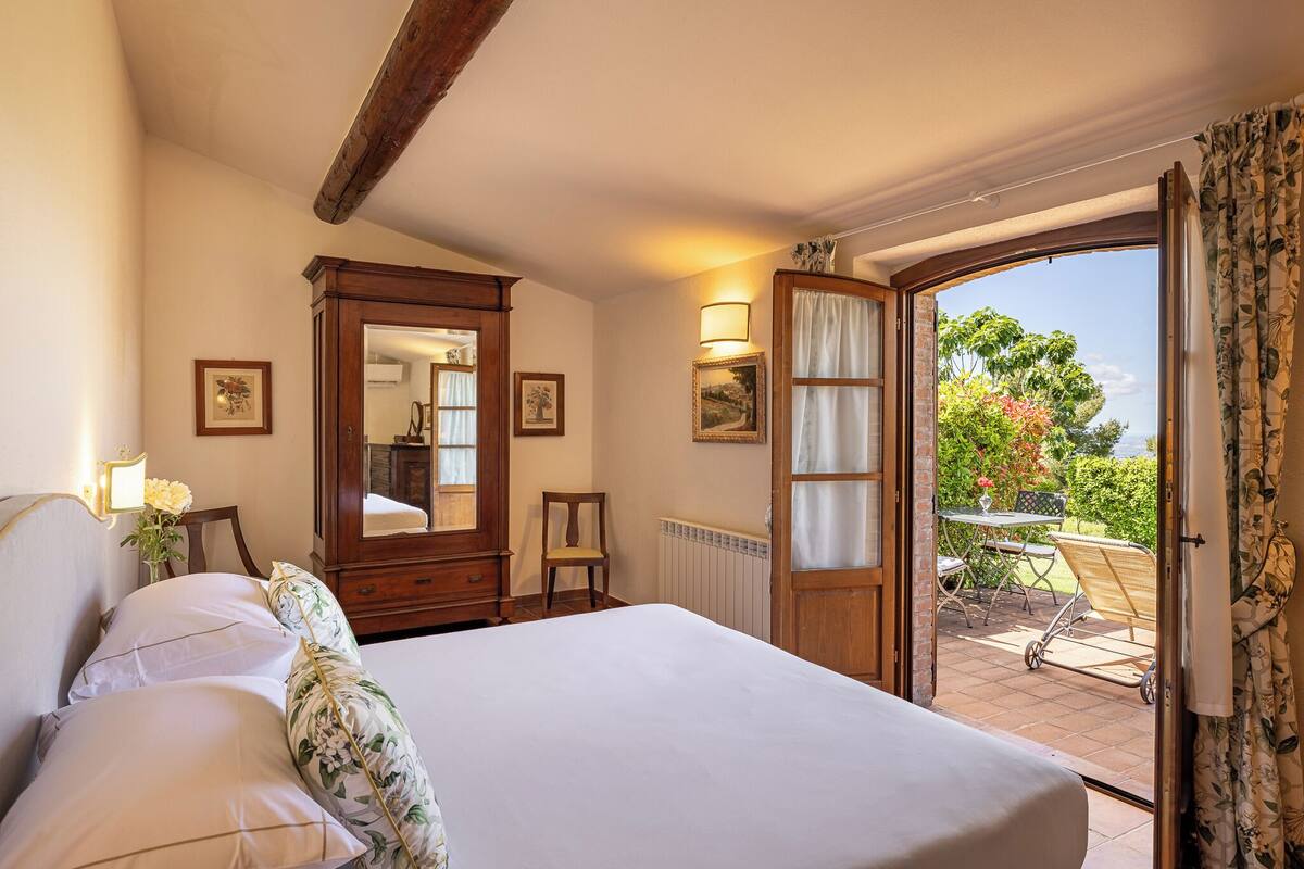 Een slaapkamer met een groot bed, een houten garderobe, een spiegel en uitzicht op een terras met een tafel en stoelen.
