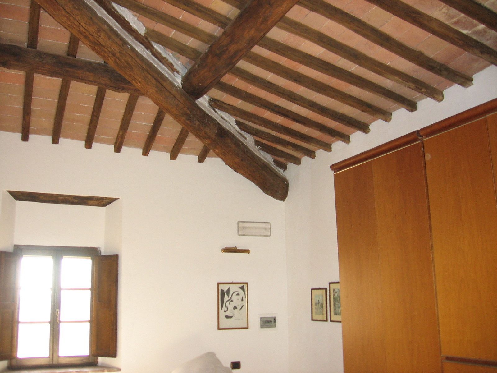 Une pièce avec un plafond en pente, des poutres en bois, une fenêtre avec des volets et une armoire.