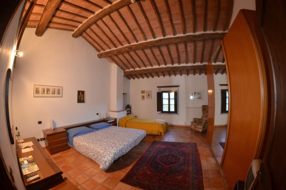 A bedroom with a wooden ceiling, a bed with a patterned bedspread, a yellow blanket, a red rug, and a wooden nightstand with books.