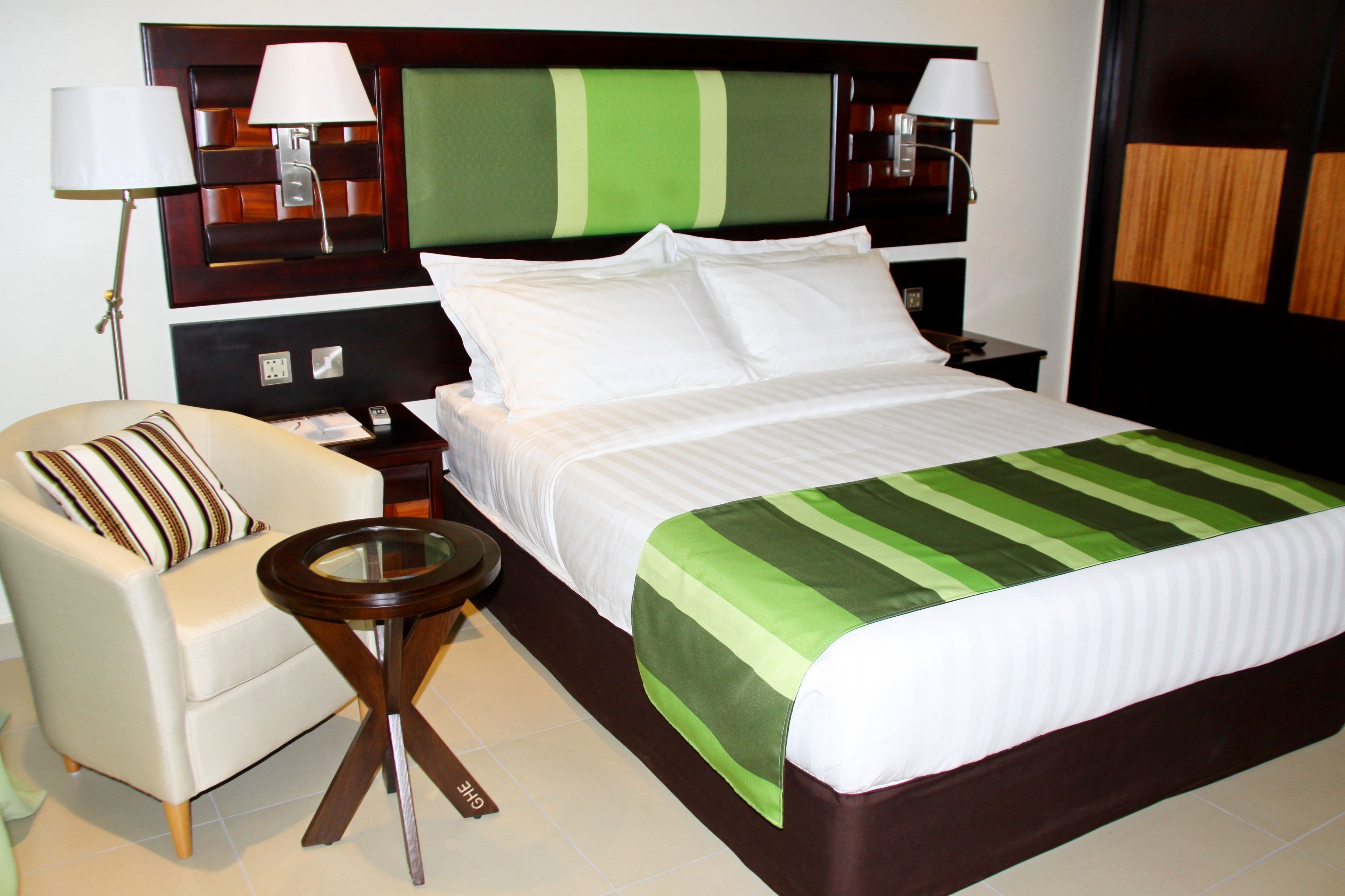 A hotel room with a bed, a chair, a small table, and two lamps.