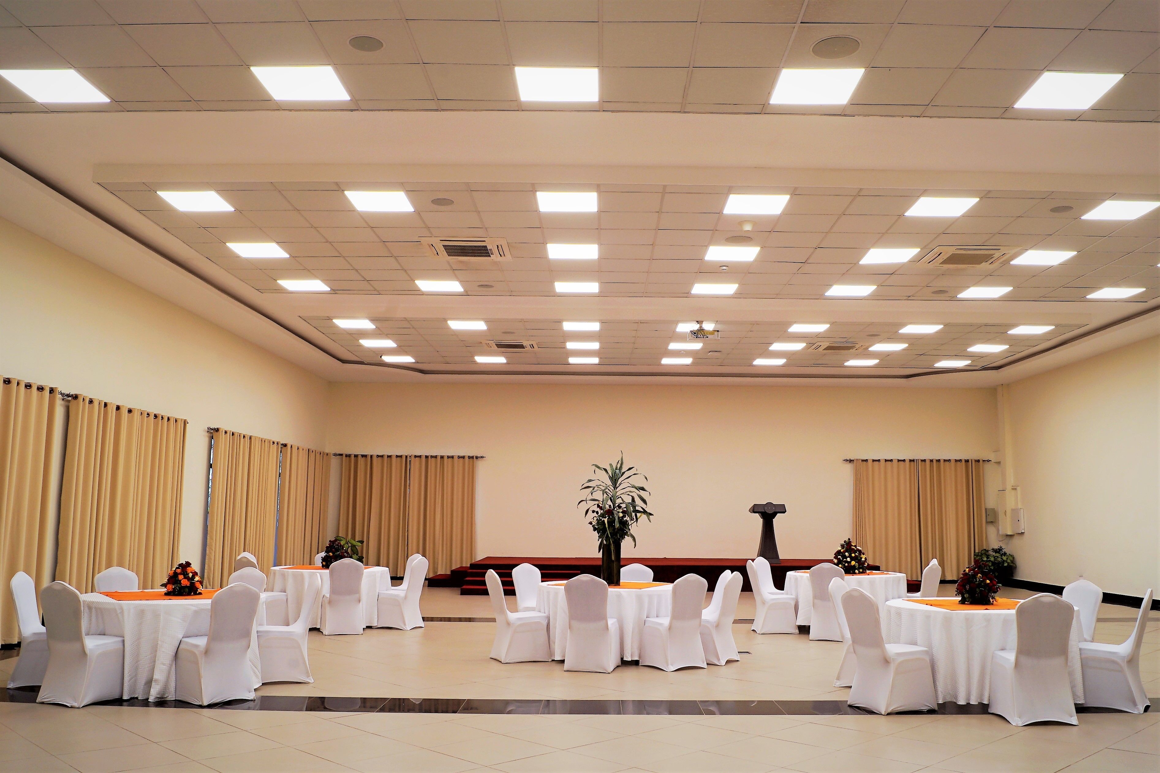 Un salón de conferencias con mesas redondas, sillas blancas y cortinas color beige.