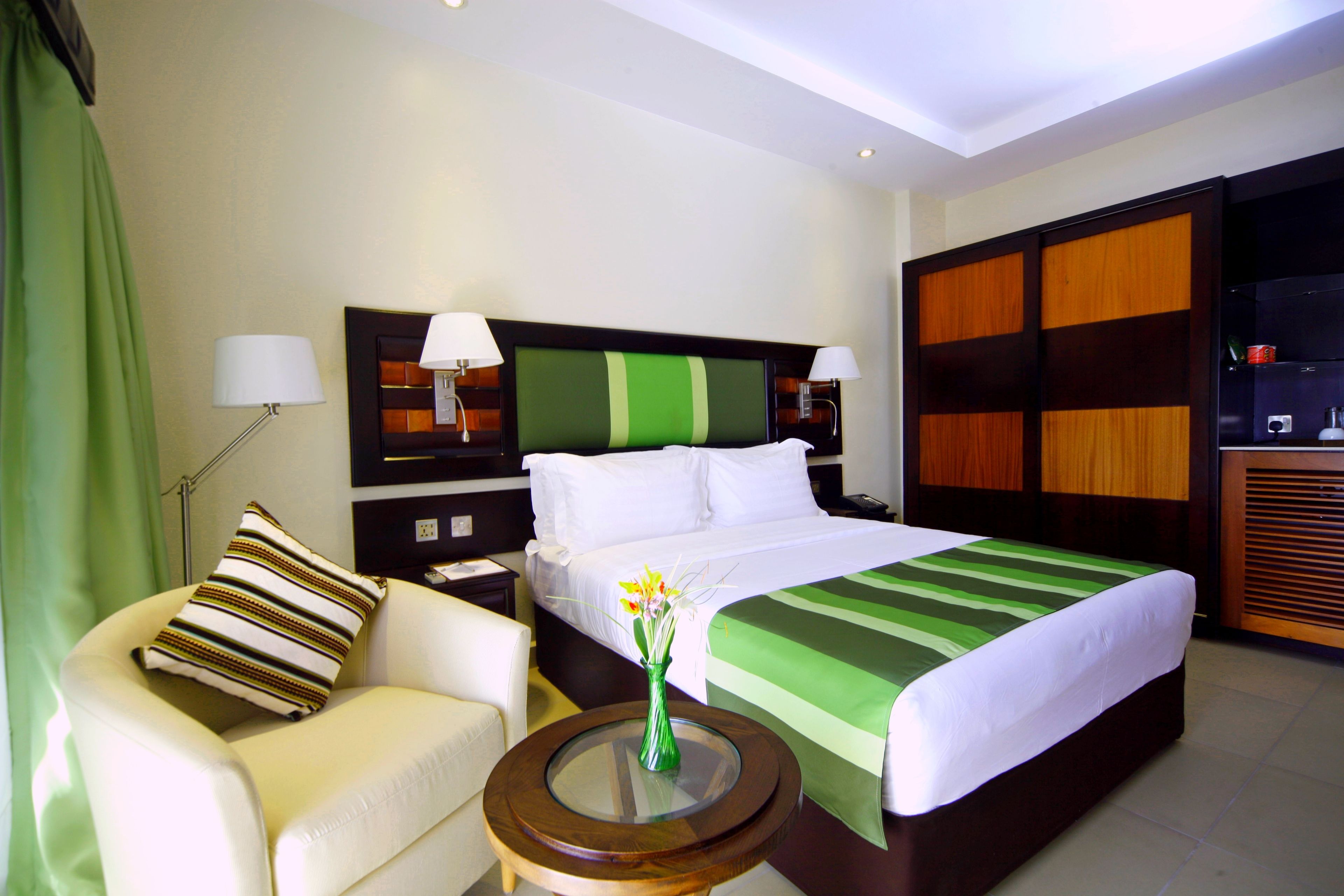 Hotelkamer met een groot bed, een groen gestreept beddegoed, een witte stoel, een klein rond tafeltje met een bloemenvaas, en een groene gordijn.
