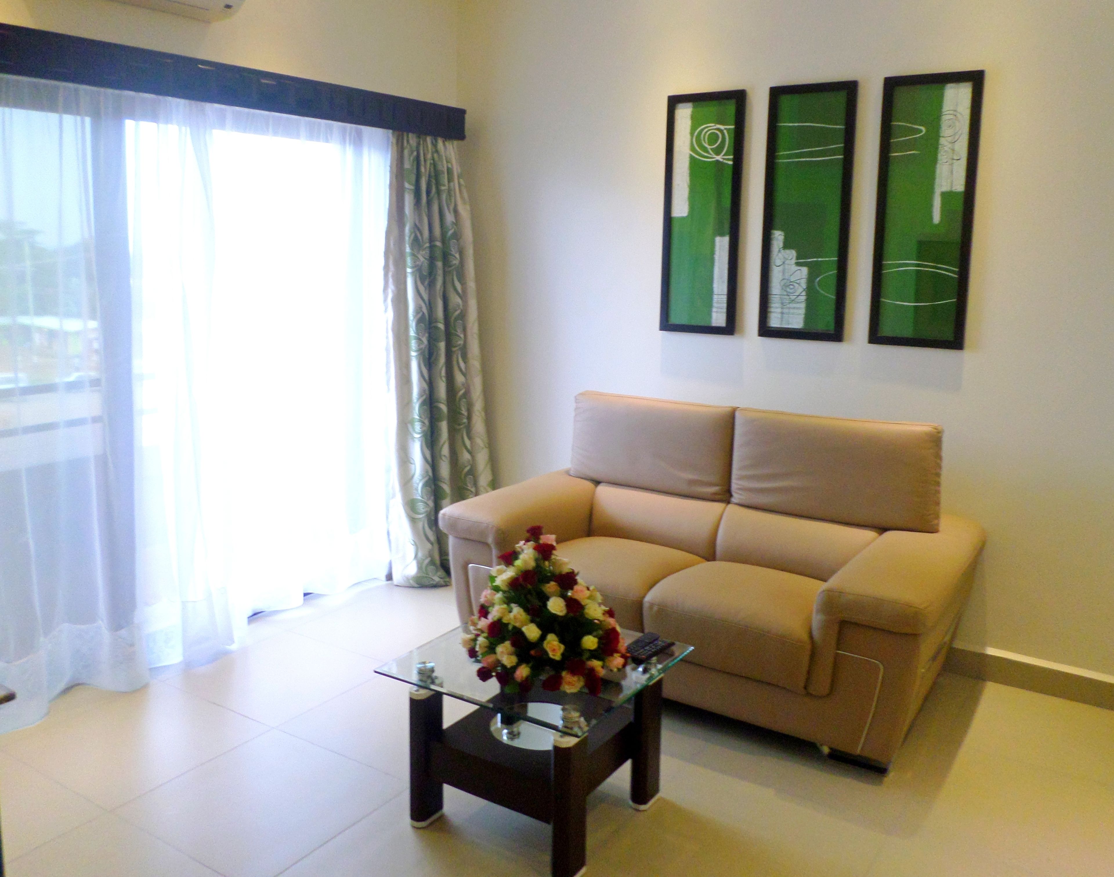 A living room with a beige sofa, a glass coffee table with a floral arrangement, and a large window with sheer curtains. There are three framed artworks on the wall.