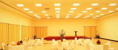 Un salón de conferencias con varias mesas redondas, sillas blancas y un escenario con un atril.