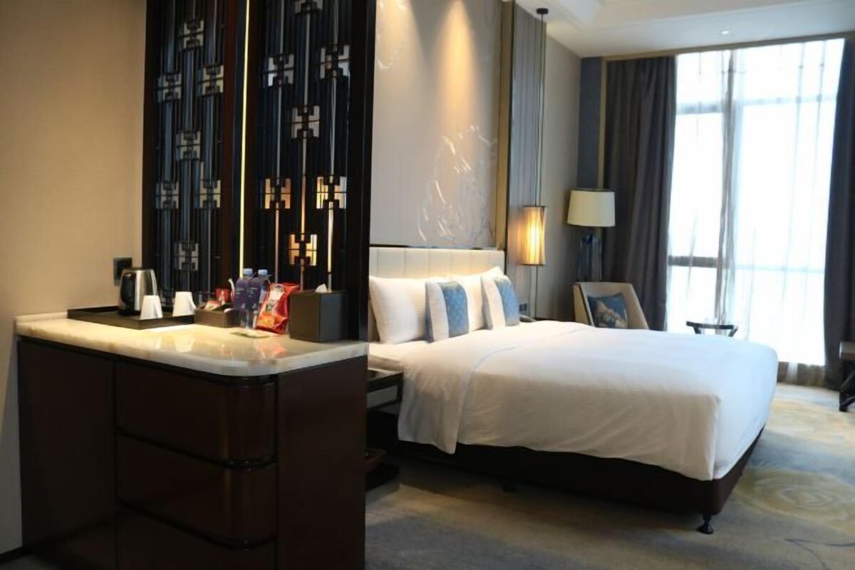 A hotel room with a bed, bedside table, lamp, and a view of the outdoors.