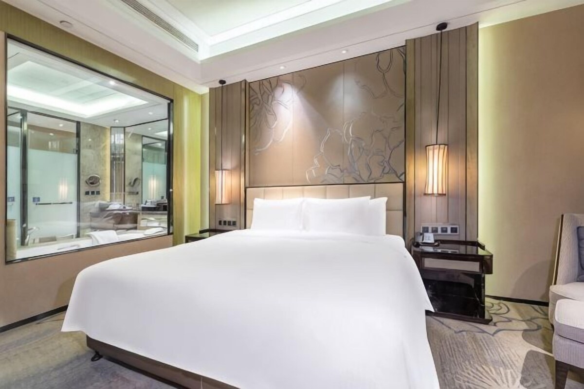 A hotel room with a large bed, a bedside table, a nightstand, a mirror, and a wall-mounted light fixture.