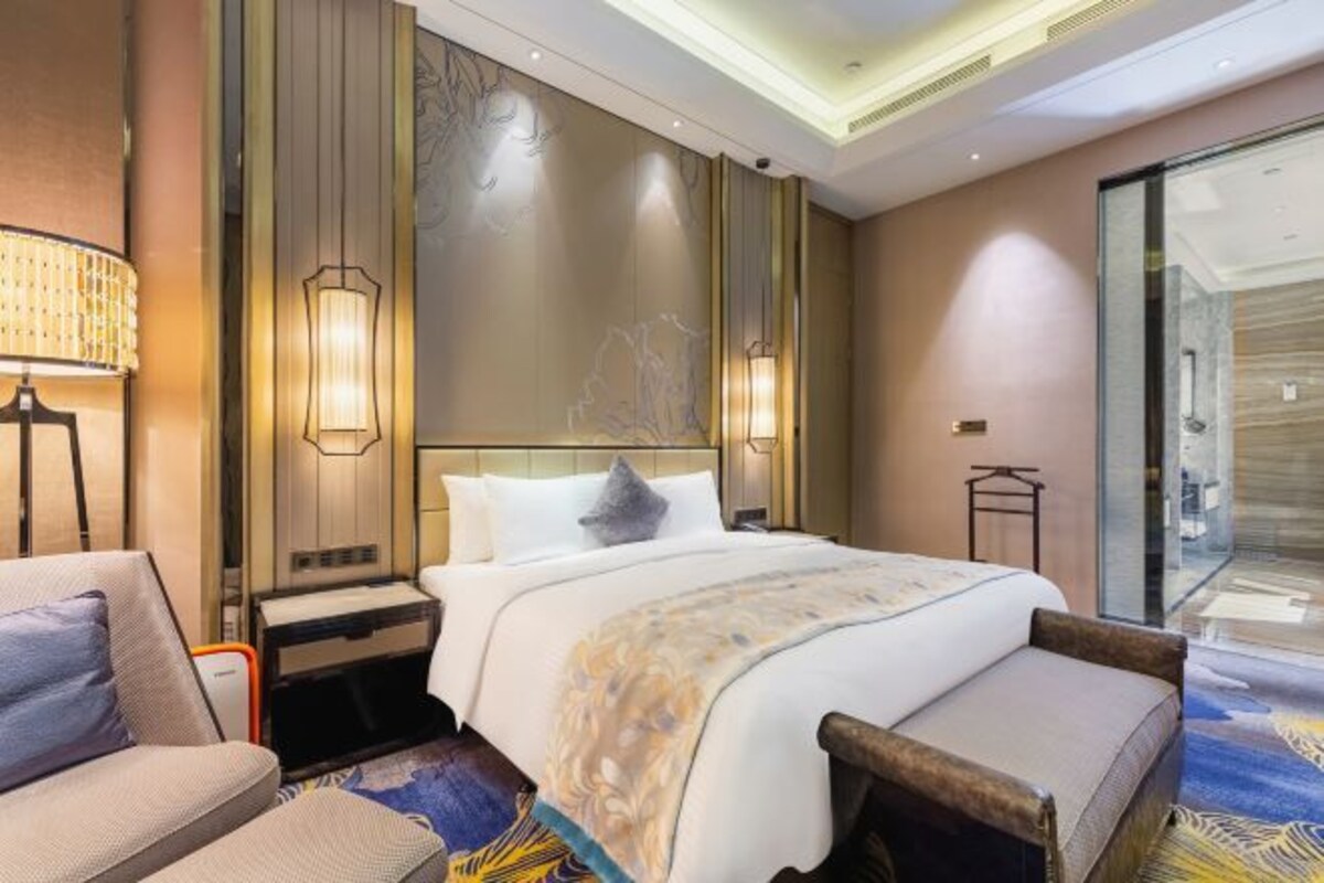 A hotel room with a bed, a chair, a nightstand, a lamp, and a wall with a decorative panel.