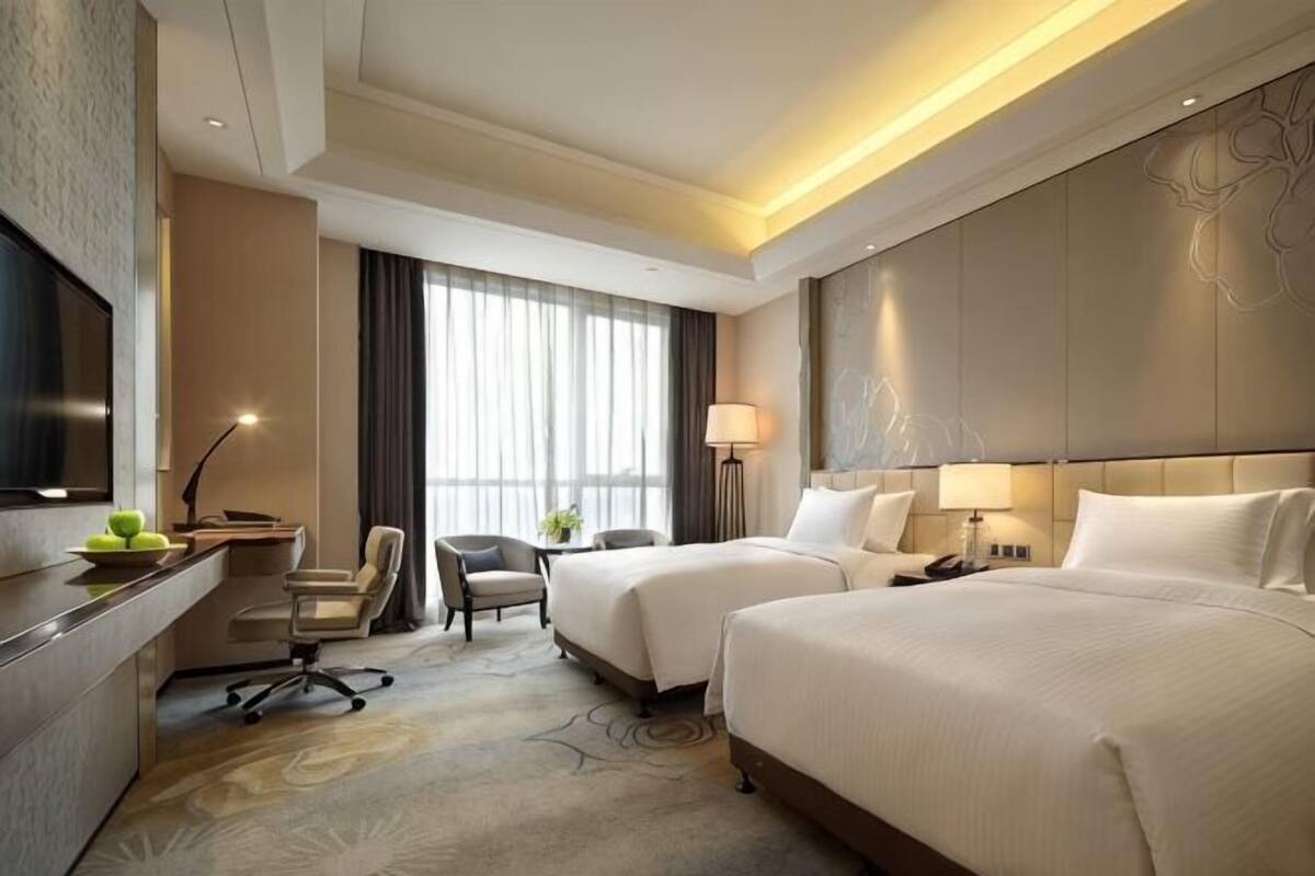 A hotel room with two beds, a desk with a chair, a TV, and a large window with curtains.