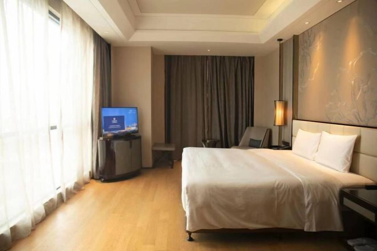 A hotel room with a large bed, a television on a stand, a desk with a chair, and a large window with curtains.