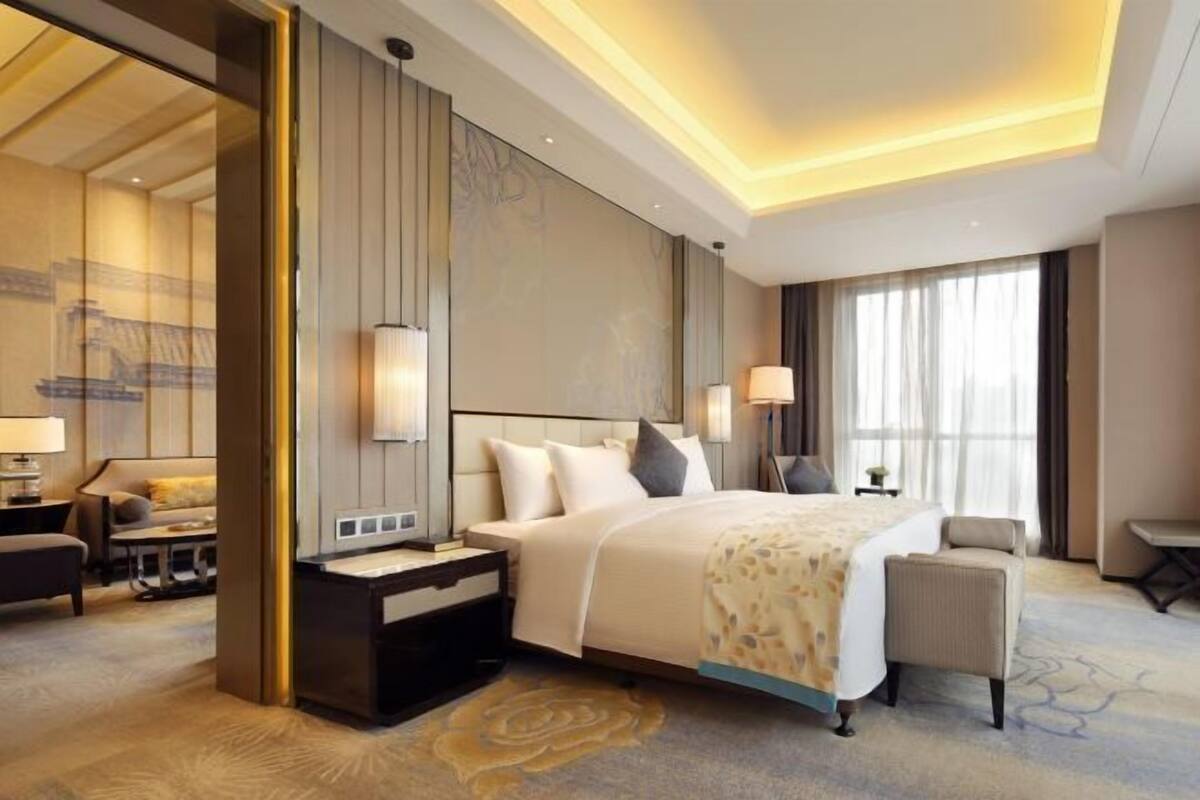 A hotel room with a bed, bedside tables, a desk, a chair, and a large window with curtains.