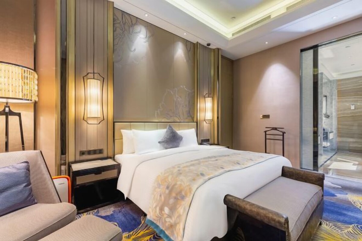 A hotel room with a bed, a chair, a nightstand, a lamp, and a wall with a decorative panel.