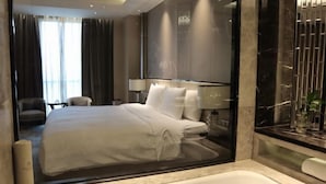 A modern hotel room with a large bed, a glass partition, a bathtub, and a view of the cityscape. - Wanda Realm Fuyang (Fuyang)