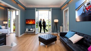 A modern hotel room with a large window, a flat-screen TV, a black leather sofa, a wooden floor, and a ceiling with decorative molding. - The Boutique Apartments (Barcelona)