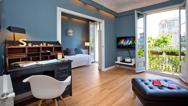 A modern bedroom with a bed, a desk with a computer, a chair, a TV, and a balcony with a view of buildings and greenery. - The Boutique Apartments (Barcelona)