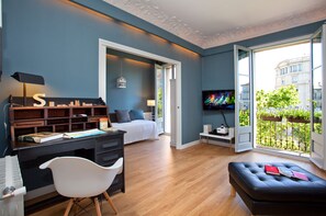 Un dormitorio moderno con una cama, un escritorio con ordenador, una silla, un televisor y un balcón con vistas a edificios y zonas verdes.