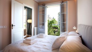 A bedroom with a large bed, an open door leading to a balcony, and a view of buildings and greenery outside. - The Boutique Apartments (Barcelona)