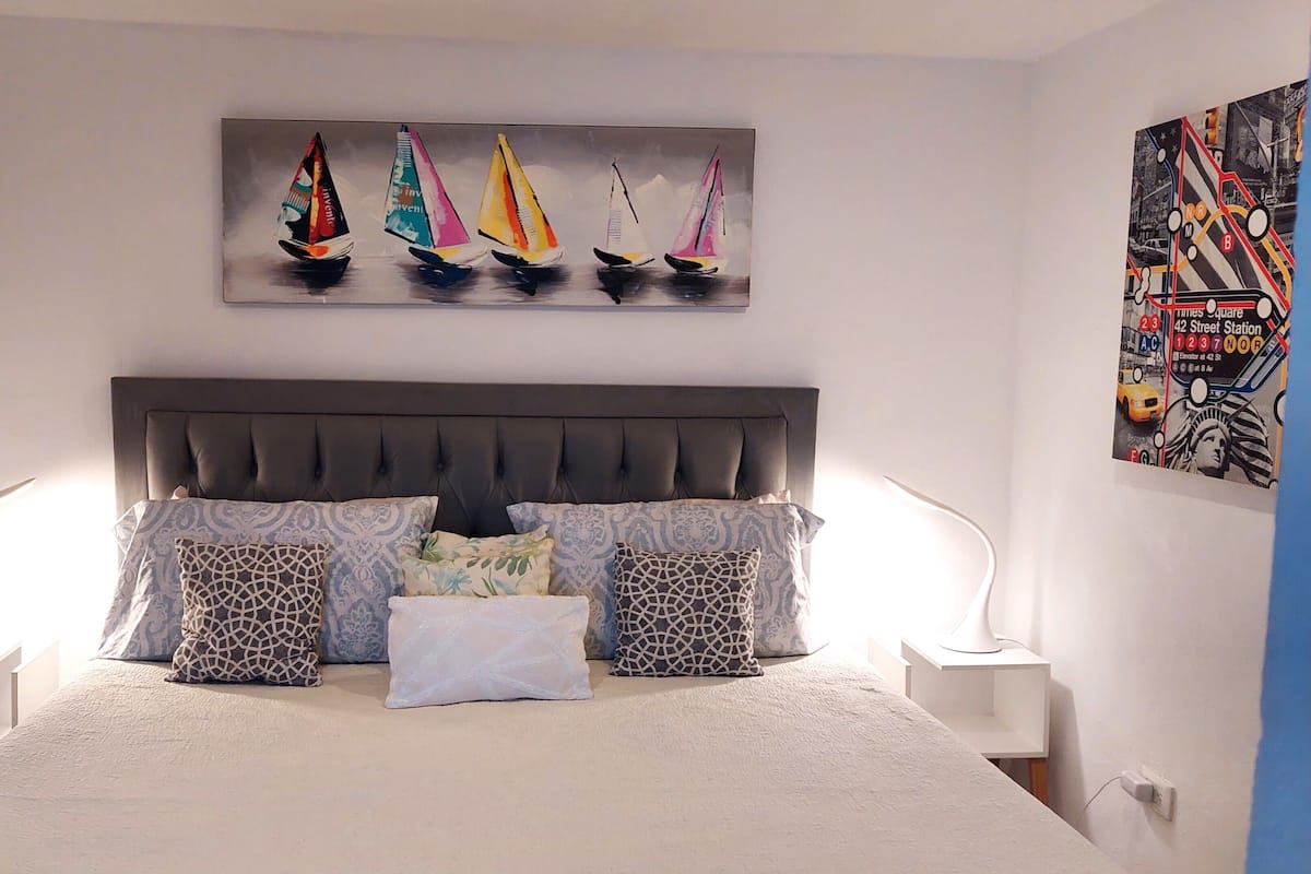 Ein Schlafzimmer mit einem großen Bett, mehreren Kissen, einem Kopfteil, Nachttischlampen und einer Wand mit einem Gemälde von Segelbooten.
