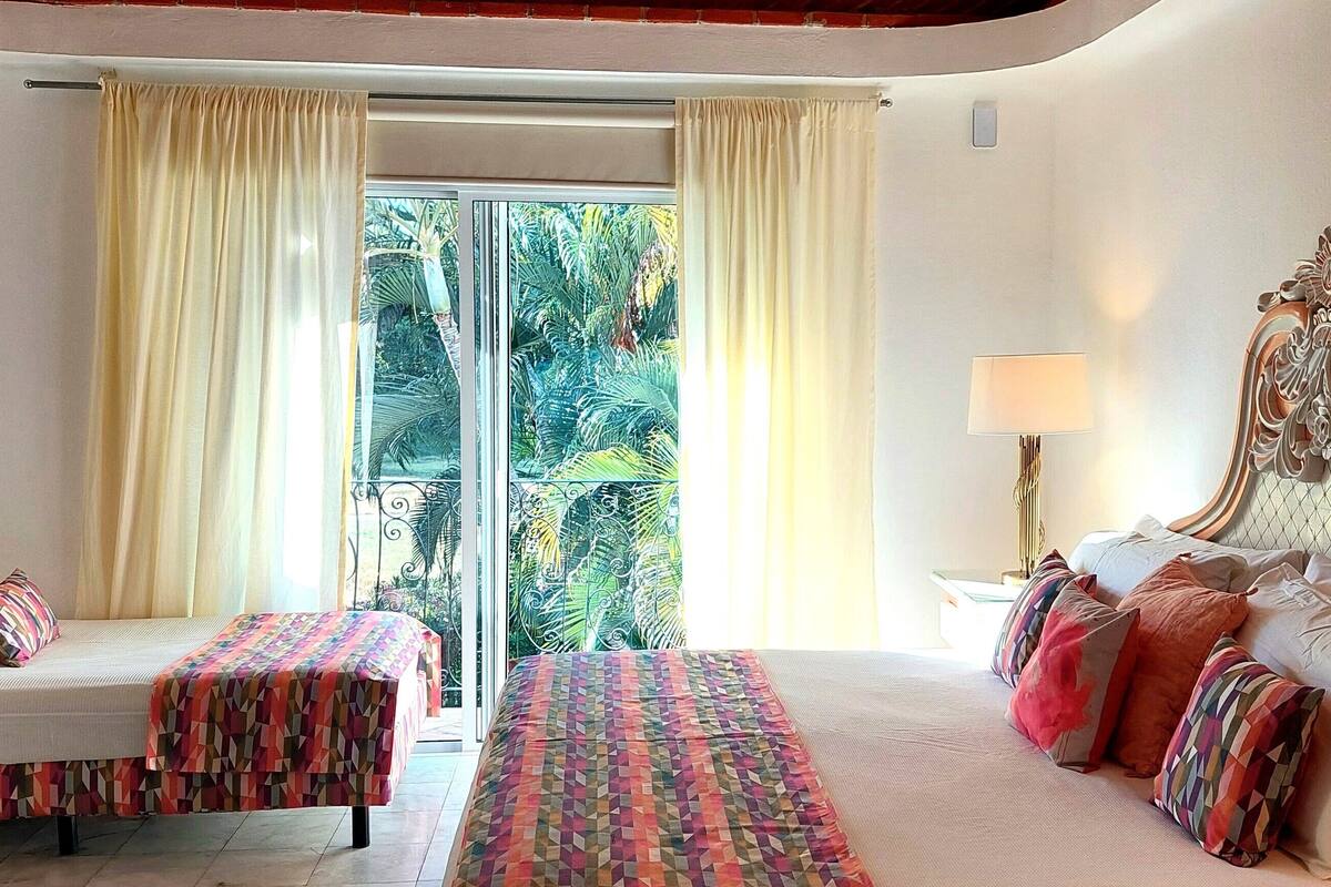 Ein Hotelzimmer mit zwei Betten, einem großen Fenster mit Vorhängen, einem dekorativen Kopfteil und einem Nachttisch mit Lampe.