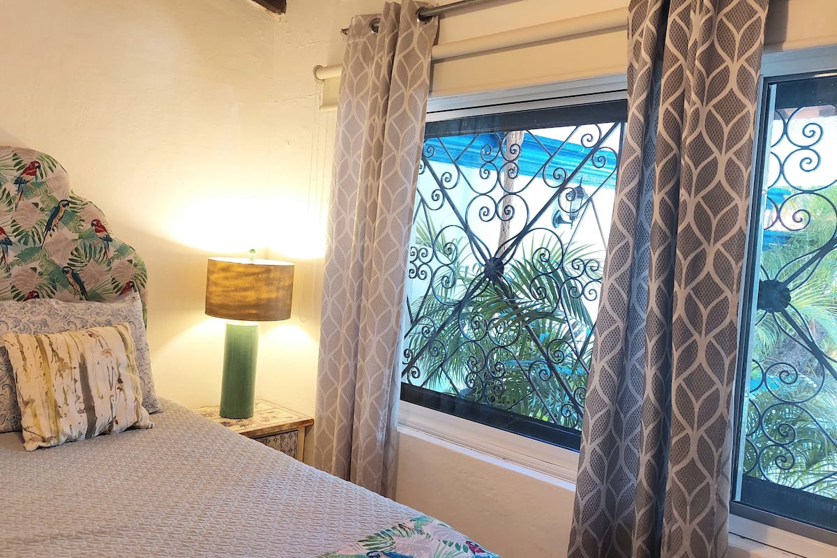 Ein Schlafzimmer mit Bett, gemustertem Kopfteil, dekorativen Kissen, einer Nachttischlampe und einem Fenster mit Vorhängen.