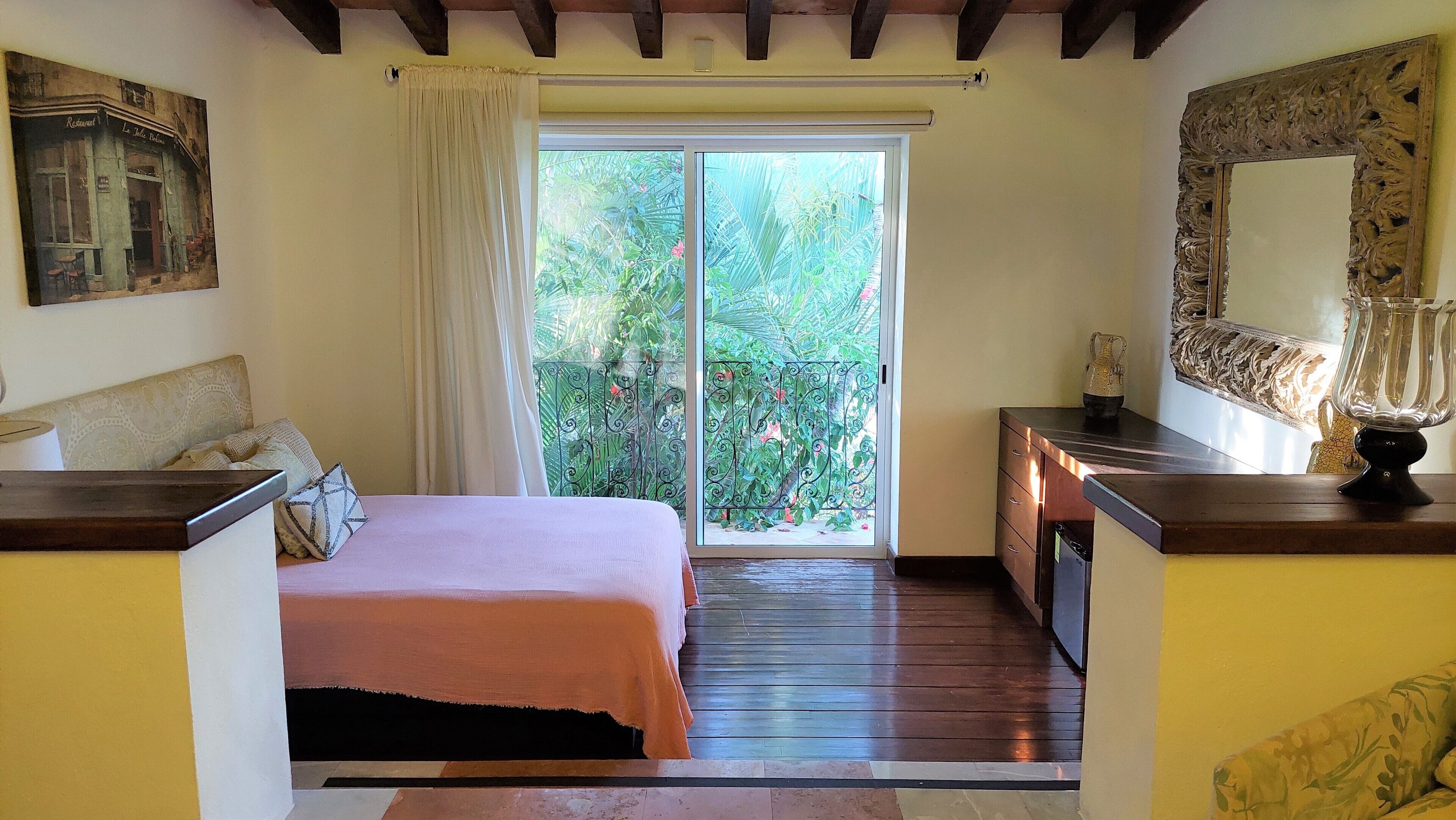 A bedroom with a bed, a small desk, a mirror, and a view of greenery through a sliding door.