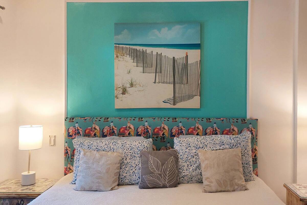 Ein Schlafzimmer mit einem Bett, zwei Nachttischlampen, einer türkisblauen Wand und einem Strandbild über dem Bett.