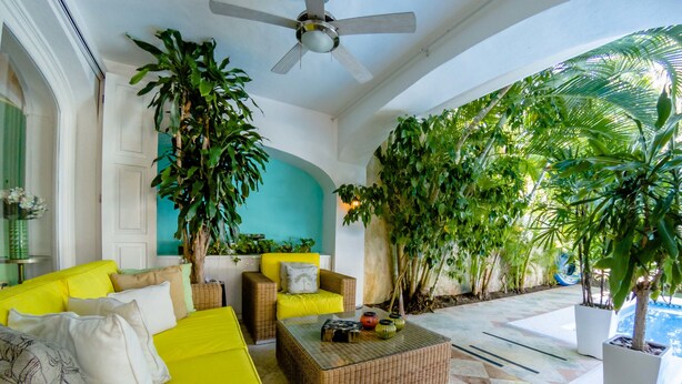Ein heller, sonnendurchfluteter Innenraum mit einem gelben Sofa, Korbmöbeln und einem Deckenventilator.