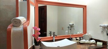 Un baño con un espejo grande, un lavamanos, dispensador de toallas, una orquídea en maceta y un secador de pelo.