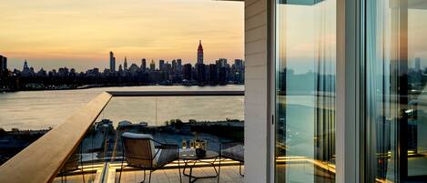 Un balcón con una mesa para dos, con vistas a un horizonte urbano y una masa de agua al atardecer.