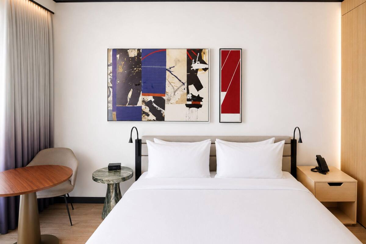 A hotel room with a bed, a bedside table, a chair, a small table, and abstract art on the wall.