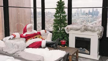 A cozy room with a fireplace, a Christmas tree, and a large window overlooking a cityscape.