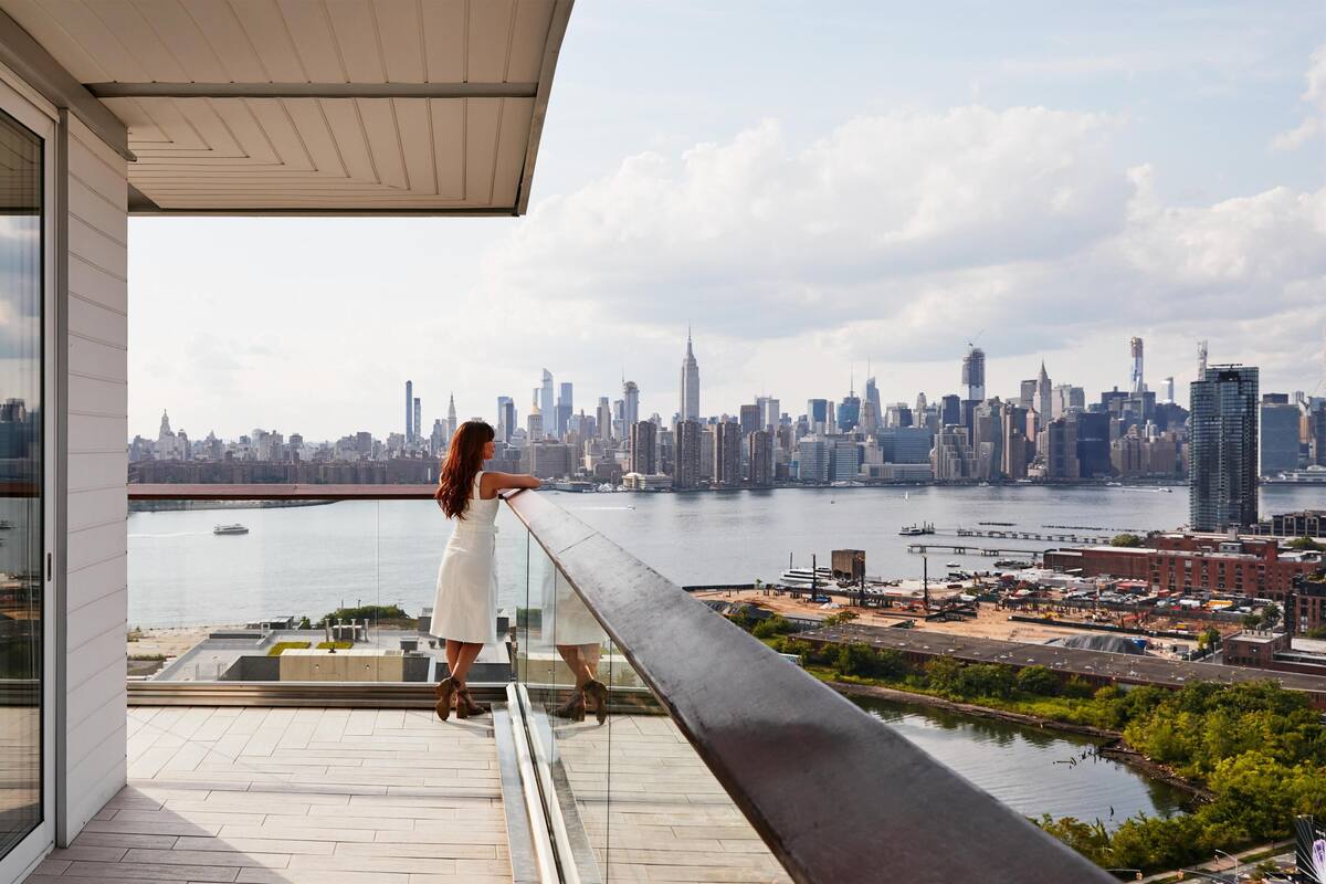 A woman standing on a balcony overlooking a city skyline with a river and buildings.